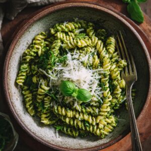 Overhead shot of a white pan on a marble surface, thick bright-green pesto sauce with diced garlic being stirred by a wooden spatula, hands visible gripping the spatula and pan handle.