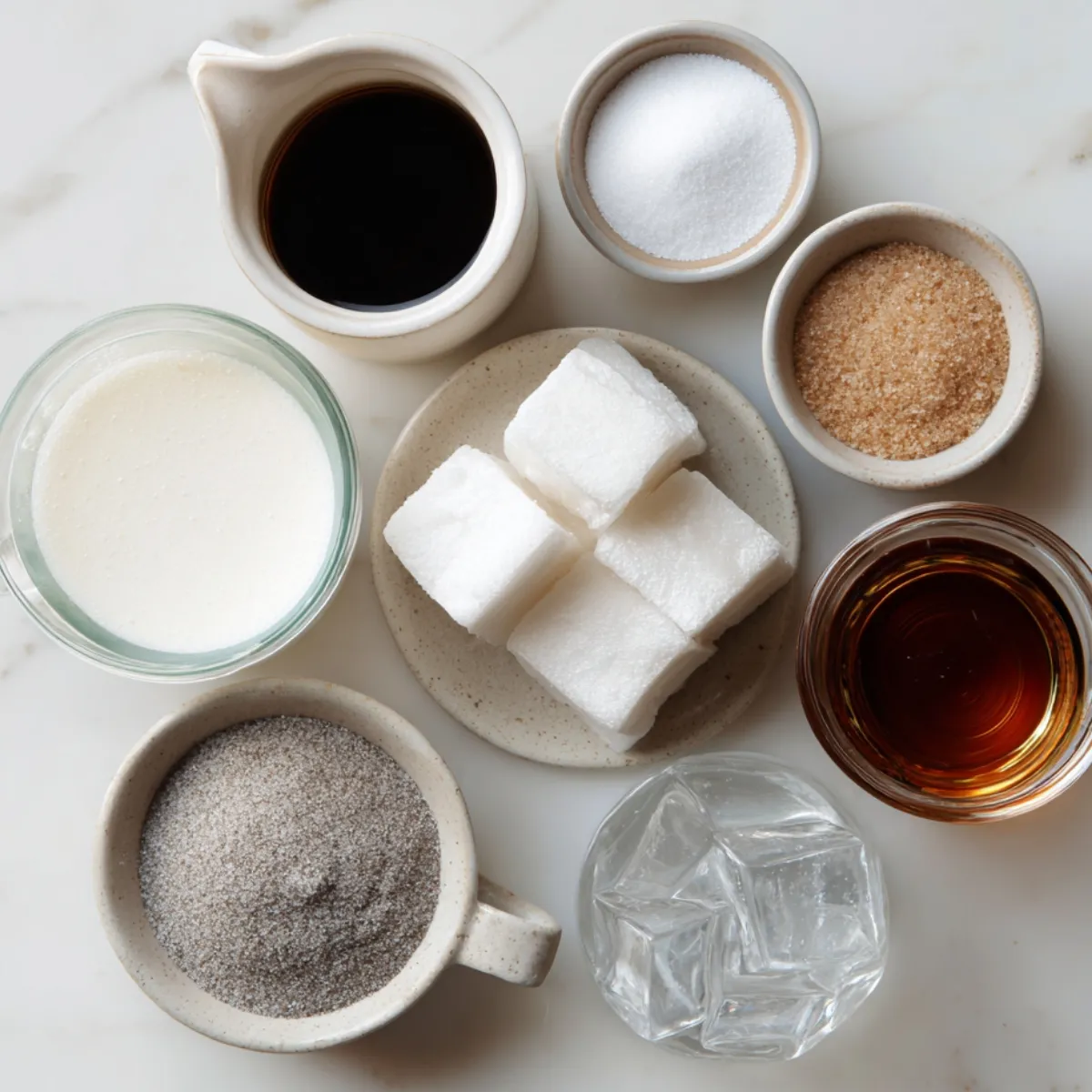 Top‑down view of matcha latte ingredients on a light marble surface: a small pitcher of hot water, bowls of white and brown sugar, a dish of honey, a cup of green matcha powder, a mug of milk, and a glass filled with ice cubes.