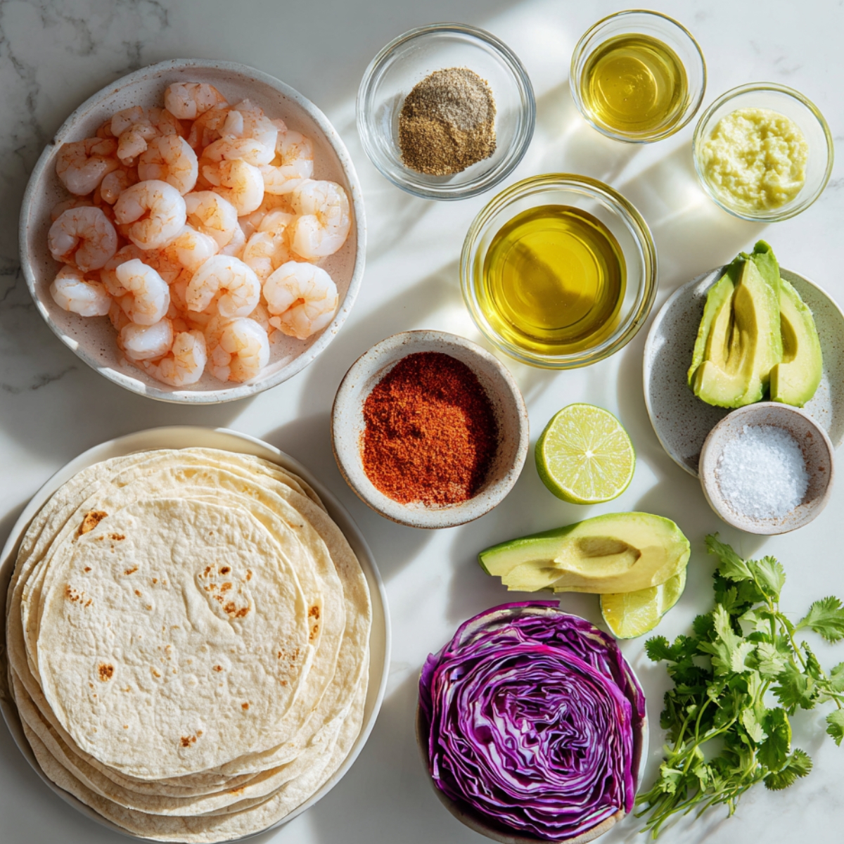 A white marble countertop with a speckled bowl of large raw shrimp; small glass bowls of olive oil, lime juice, and minced garlic; ramekins of chili powder, cumin, paprika, and salt & pepper; a stack of corn and flour tortillas; sliced avocado and lime halves; shredded red cabbage; and a bunch of fresh cilantro.