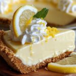 A creamy slice of homemade no-bake lemon pie with a golden graham cracker crust, topped with whipped cream, a lemon slice, mint leaf, and lemon zest. Served on a wooden board with a lemon wedge, the whole pie is softly visible in the background.