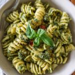 A rustic bowl of fusilli pesto pasta tossed in bright green basil pesto, topped with a sun-dried tomato cluster and fresh basil sprig, with a fork resting on the rim.