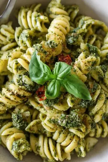 A rustic bowl of fusilli pesto pasta tossed in bright green basil pesto, topped with a sun-dried tomato cluster and fresh basil sprig, with a fork resting on the rim.