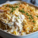 A white ceramic baking dish of homemade poppy seed chicken casserole with a golden breadcrumb-and-cheddar crust, black poppy seeds, and parsley, a wooden spoon lifting cheesy chicken and pasta strands.
