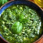 A bowl of vibrant green Basil Pesto Recipe topped with a basil leaf, with fresh basil and olive oil on the side