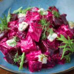 A blue plate filled with a vibrant beetroot salad, featuring creamy beet chunks, crumbles of cheese, fresh dill, and arugula leaves as garnish.