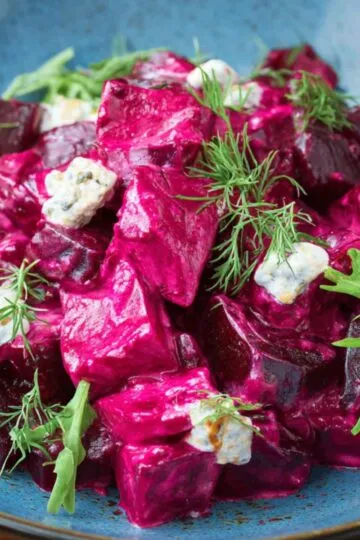 A blue plate filled with a vibrant beetroot salad, featuring creamy beet chunks, crumbles of cheese, fresh dill, and arugula leaves as garnish.
