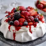 Elegant Berry Pavlova dessert with a crisp meringue base, whipped cream topping, and a mix of fresh strawberries, raspberries, blackberries, and blueberries, drizzled with berry sauce, served on a black cake stand.