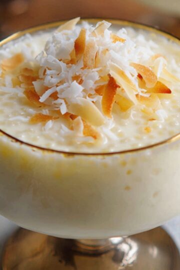 Glass dessert cups of coconut tapioca pudding, topped with toasted coconut flakes, set on a light blue surface with spoons in the background.