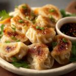 Golden brown, crispy-bottomed Fried Dumplings served on a ceramic plate. Topped with chopped scallions and paired with a small bowl of soy-based dipping sauce. Fresh cilantro garnishes the side. These are pan-fried dumplings with perfect texture contrast – crispy and juicy.