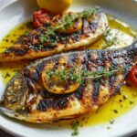 Two grilled fish on a plate with lemon slices, fresh thyme, and tomatoes, served in a golden olive oil sauce.