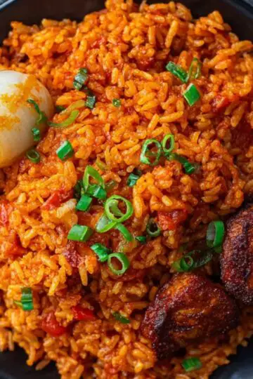 A black plate of Jollof rice garnished with chopped scallions, served with caramelized onions and fried plantains, placed on a dark napkin next to a fork.