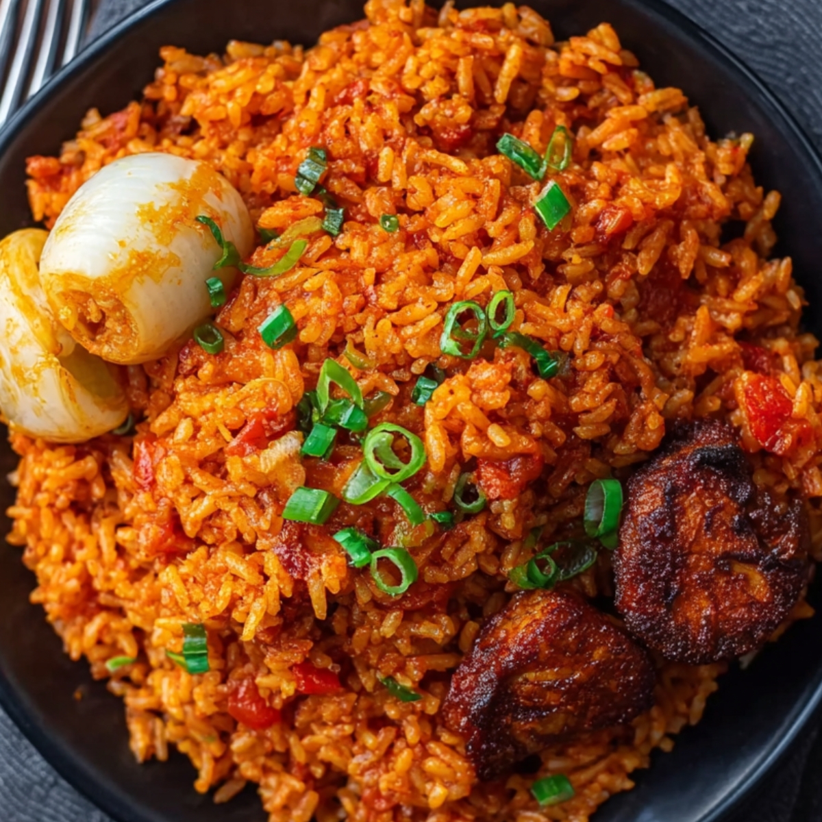 A black plate of Jollof rice garnished with chopped scallions, served with caramelized onions and fried plantains, placed on a dark napkin next to a fork.