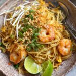 Bowl of Pad Thai with shrimp, bean sprouts, crushed peanuts, lime wedges, and green onions, served on a rustic ceramic plate with chopsticks and a fork.