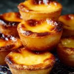 A plate stacked with golden-brown Portuguese Custard Tarts , their tops caramelized and blistered, on a decorative blue and white plate.