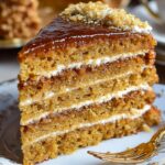 A close-up of a slice of traditional Russian honey cake recipe(Medovik) served on a vintage floral porcelain plate with a gold fork. The cake features multiple thin honey layers filled with creamy sour cream frosting and topped with a glossy caramel glaze and crushed nuts.