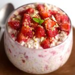 A serving of strawberry cheesecake overnight oats in a clear glass cup, topped with fresh diced strawberries and a mint leaf, with a spoon beside it.