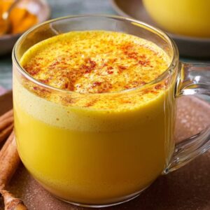 A glass mug filled with frothy Turmeric Golden Milk, topped with cinnamon and surrounded by cinnamon sticks on a brown plate