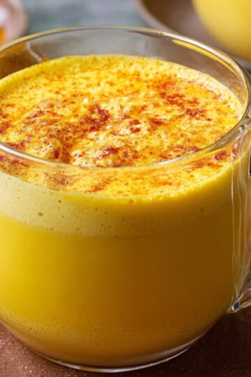 A glass mug filled with frothy Turmeric Golden Milk, topped with cinnamon and surrounded by cinnamon sticks on a brown plate