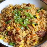 A wooden bowl of pork fried rice recipe : golden fried rice with scrambled egg, green peas, diced carrots, and crispy pork pieces, garnished with sliced green onions and herbs on top.