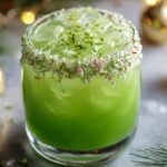 A Grinch Punch Recipe in a glass rimmed with crushed peppermint and lime zest, decorated with holiday ornaments and pine branches.