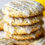 A close-up of lemon poppy seed cookies stacked high, drizzled with shiny glaze, with whole lemons in the background.