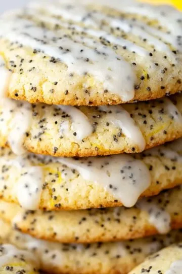 A close-up of lemon poppy seed cookies stacked high, drizzled with shiny glaze, with whole lemons in the background.
