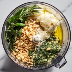 A food processor filled with basil, pine nuts, garlic, cheese, and olive oil, ready to be blended into pesto.