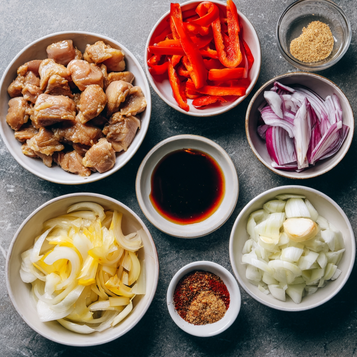 Bowls of chopped ingredients including marinated chicken, red bell peppers, soy sauce, red and white onions, garlic, and spice blends, arranged on a stone surface.