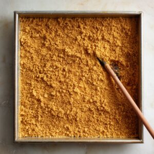 A baking pan filled with pressed graham cracker crust, smoothed with a spoon.