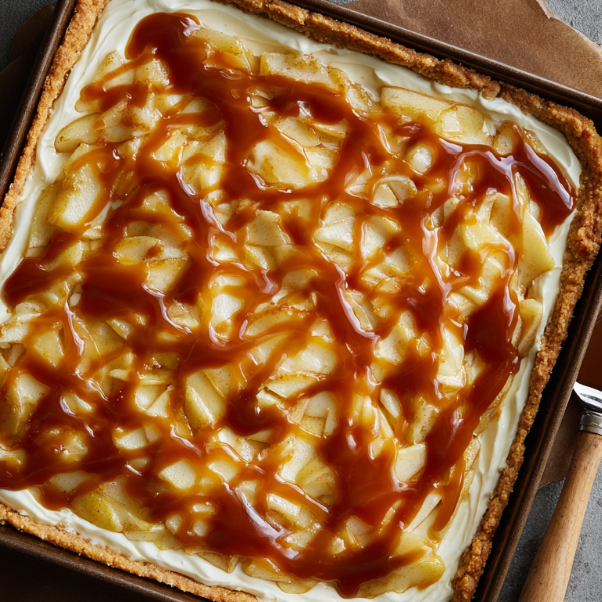 A full cheesecake bar pan topped with cooked apple slices and caramel drizzle, ready to chill.