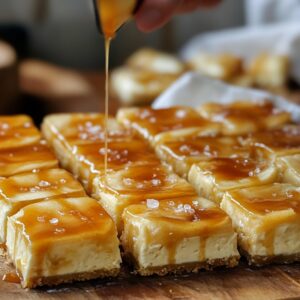 Cheesecake bars cut into neat squares on a wooden board, being drizzled with golden caramel sauce.