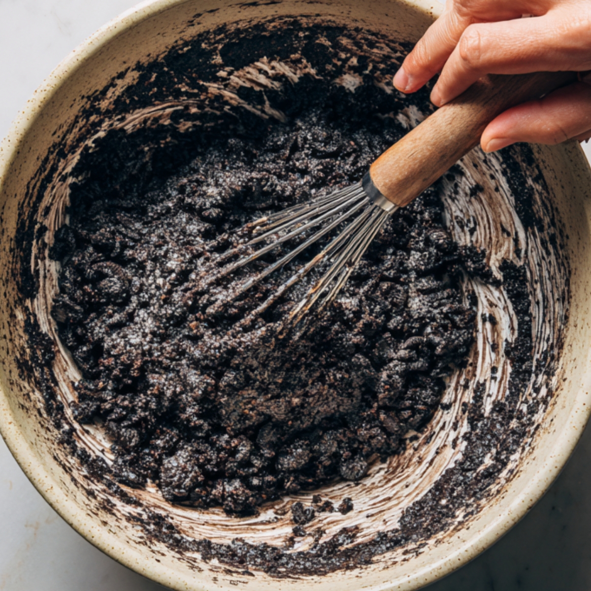 A ceramic bowl containing thick Oreo batter being mixed with a whisk, with visible crushed cookies throughout.