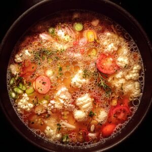 Top-down view of a bubbling pot of vegetable soup with floating slices of tomato, green onions, cauliflower florets, carrots, and herbs in a golden broth, with visible oil bubbles on the surface.