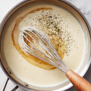 A saucepan with creamy white sauce being whisked with herbs and pepper.