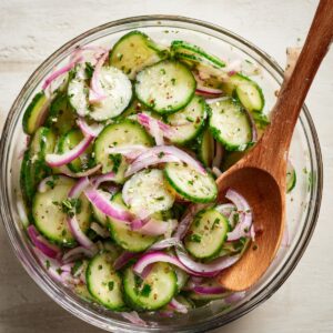 Top-down view of a cucumber and red onion salad tossed with herbs in a glass bowl, mixed with a wooden spoon.