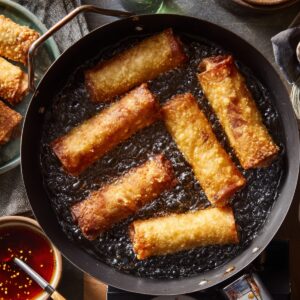 Overhead shot of egg rolls frying in bubbling hot oil inside a deep pan. Several rolls are perfectly golden, while others are still cooking. A plate of fried rolls and a bowl of chili dipping sauce sit beside the pan.