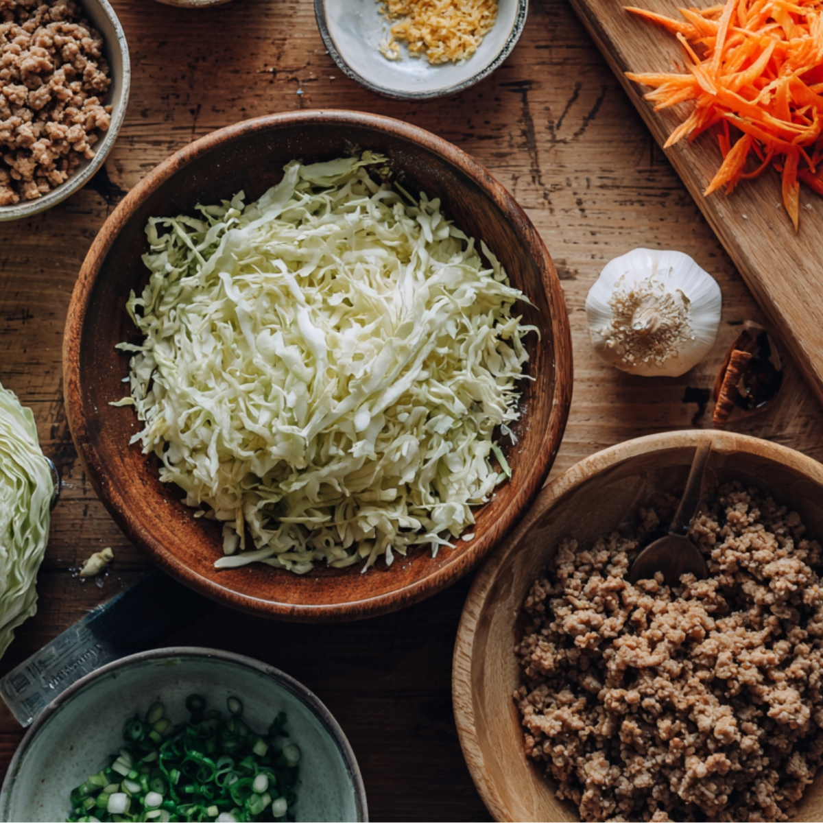 Top-down view of a rustic kitchen counter with bowls of shredded cabbage, cooked ground meat, chopped carrots, garlic, soy sauce, and sliced green onions. All ingredients are prepped for egg roll filling.
