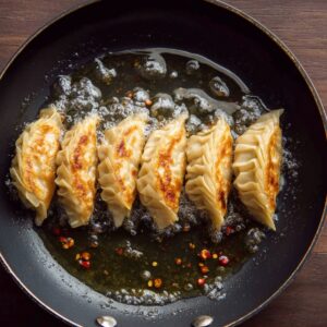 Six dumplings frying in hot oil inside a black nonstick pan. The dumplings have golden-brown bottoms and crimped edges, with chili flakes floating in the oil. The background is a dark wooden surface.