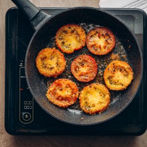 Breaded tomato slices sizzling in a frying pan with a golden crust forming on each piece.