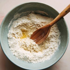 A bowl of flour and oil being combined with a wooden spoon to form dough.
