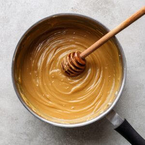 A hand pouring warm honey–peanut butter mixture over an oat and dried fruit mixture in a parchment-lined pan.