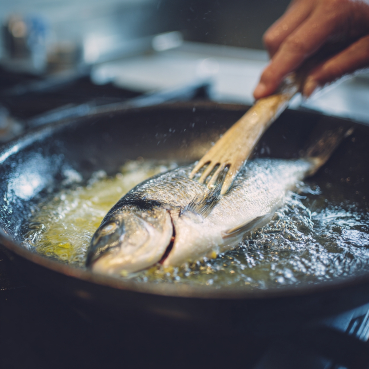 Fresh whole fish being cooked in hot oil in a frying pan, held with tongs over the bubbling surface.