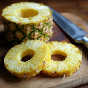 A freshly cut pineapple with slices on a wooden board, showing the cored rings and a knife beside it.
