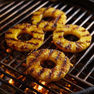 Pineapple rings cooking on a grill, showing charred grill marks over glowing flames.