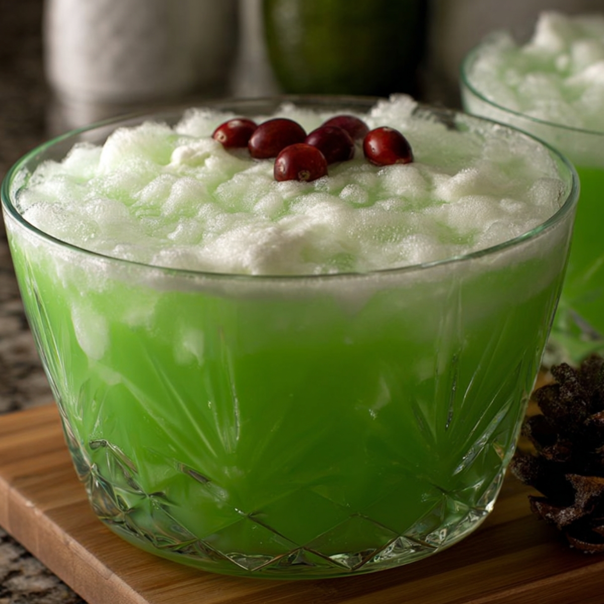 A large glass punch bowl filled with green frothy punch, topped with red cranberries, sitting on a kitchen counter.
