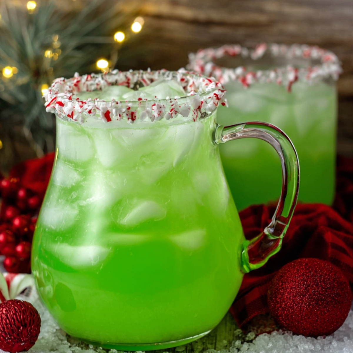 A green iced drink in a glass mug with a handle, rimmed with crushed peppermint candy, set among Christmas decorations.