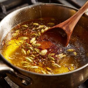 A saucepan filled with golden sugar syrup simmering with green cardamom pods and saffron strands, stirred with a wooden spoon.
