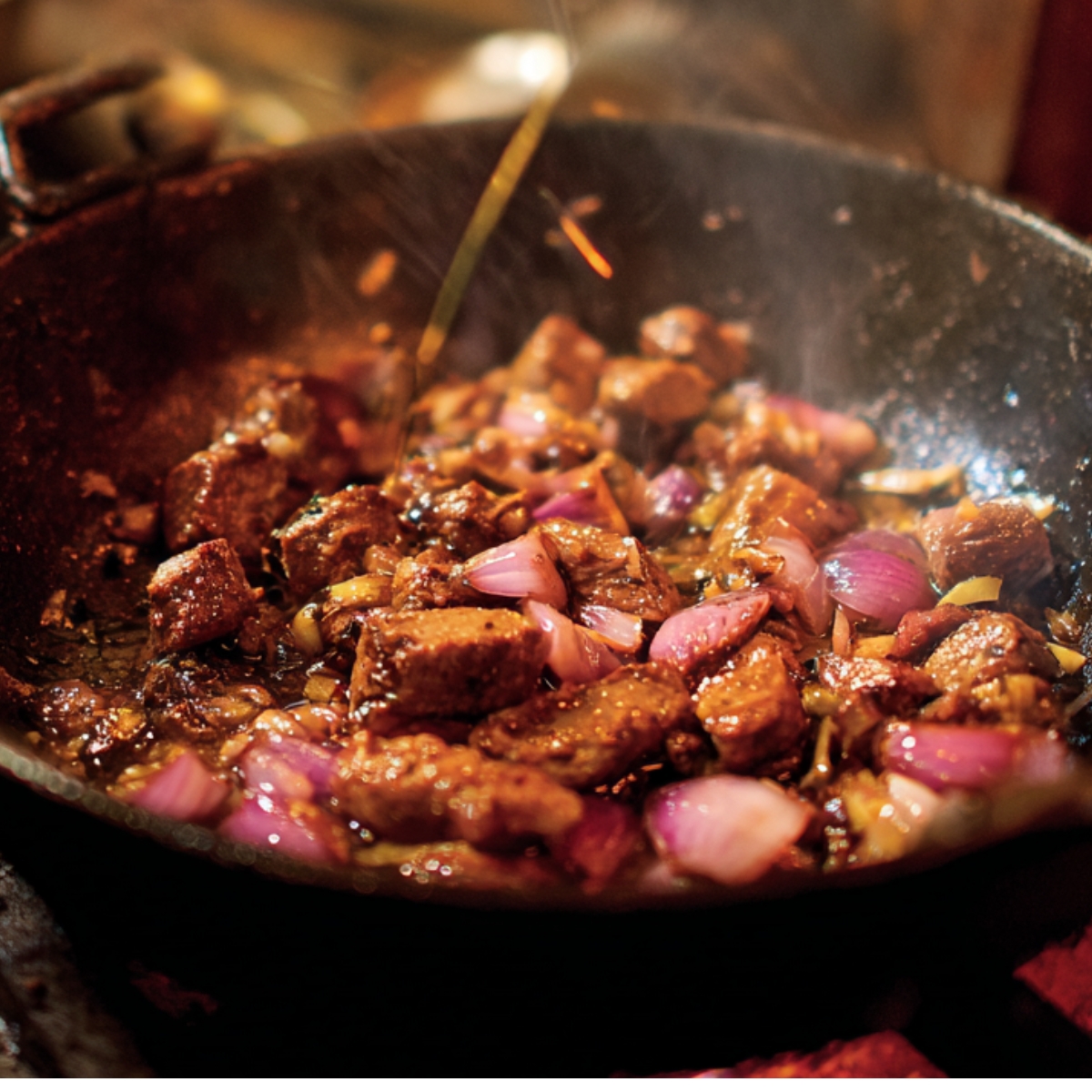 A rustic black wok with chunks of meat and chopped red onions being sautéed in hot oil over an open flame.