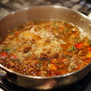 A shallow stainless steel pan with a bubbling pepper mixture of minced meat, red peppers, and herbs simmering on a gas stove.