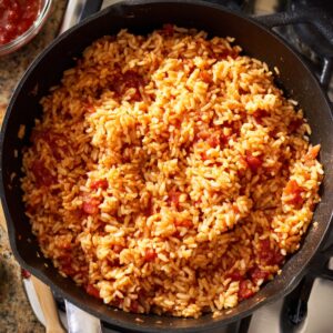 A cast iron pan filled with vibrant orange-red Jollof rice sitting on a stovetop, with a jar of sauce and wooden spoon nearby.
